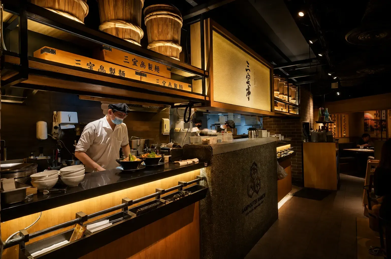 Chef in a mask preparing ramen bowls behind a warm wooden open-kitchen counter at Sanpoutei Ramen.