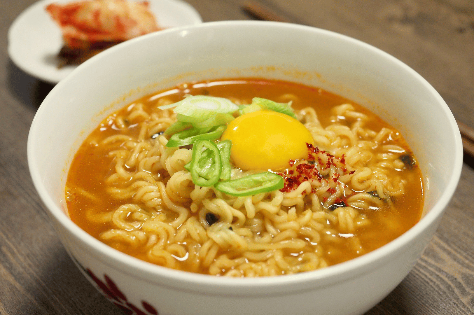 A bowl of spicy ramen in orange broth, crowned with a raw egg yolk, green onions, and red chili flakes. A side of kimchi peeks from the background, enhancing the dish’s fiery character. Set on a wooden table, the scene evokes the bold flavors and casual charm of Seoul’s noodle havens—where heat, texture, and tradition converge in every slurp.