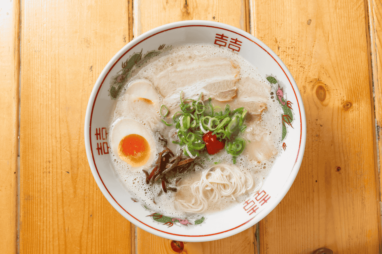 A bowl of tonkotsu ramen rests on a wooden surface, its creamy pork bone broth cradling thin noodles, tender chashu slices, a soft-boiled egg with molten yolk, and a dollop of spicy red paste. Garnished with green onions and dark mushrooms, the dish is served in a traditional bowl adorned with red Chinese characters—capturing the final, flavorful result of meticulous bone extraction and slow emulsification.