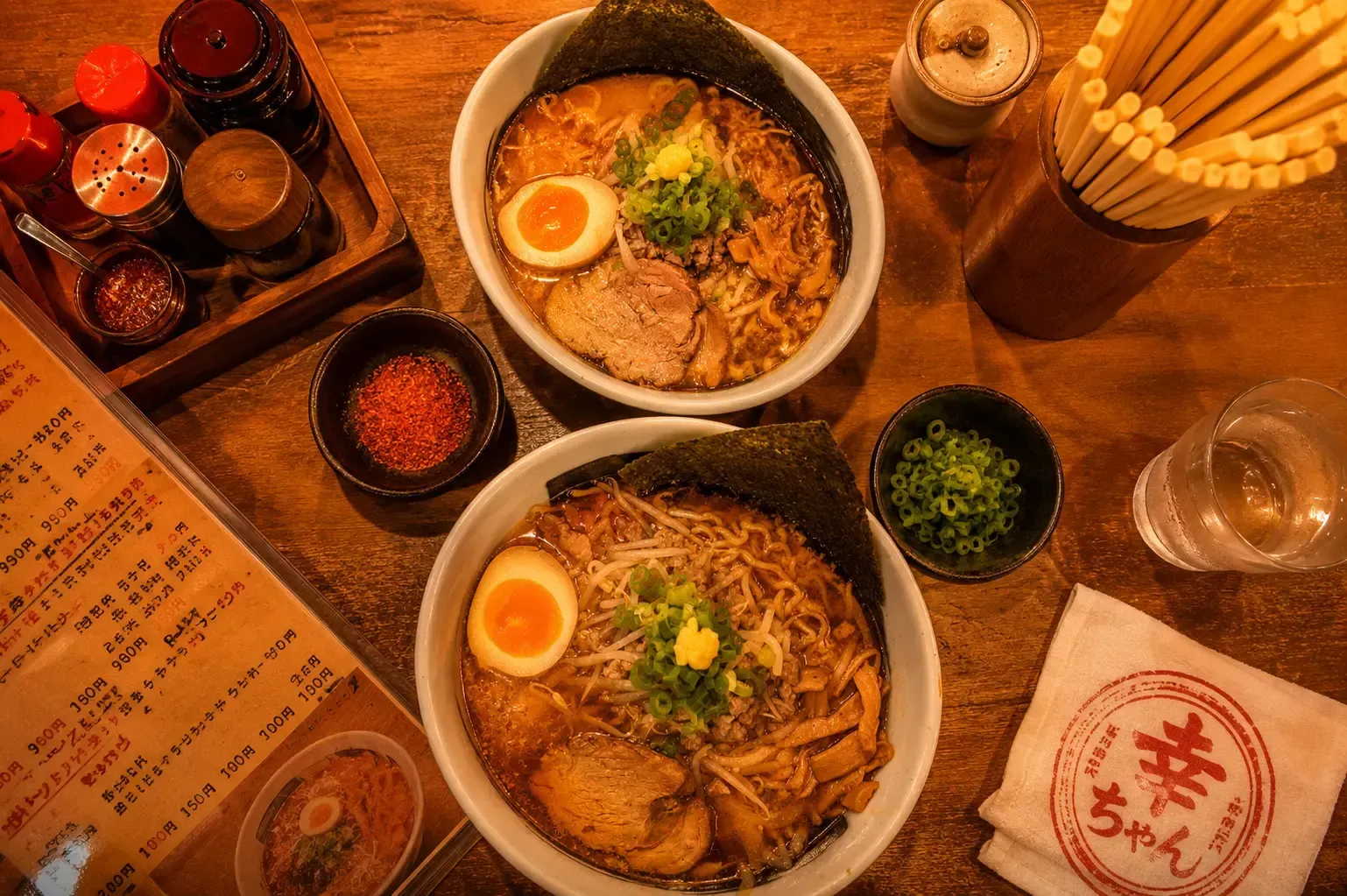 An overhead view of two bowls of ramen with chashu pork, soft-boiled egg, and nori on a wooden table, surrounded by a menu, spices, green onions, and chopsticks.