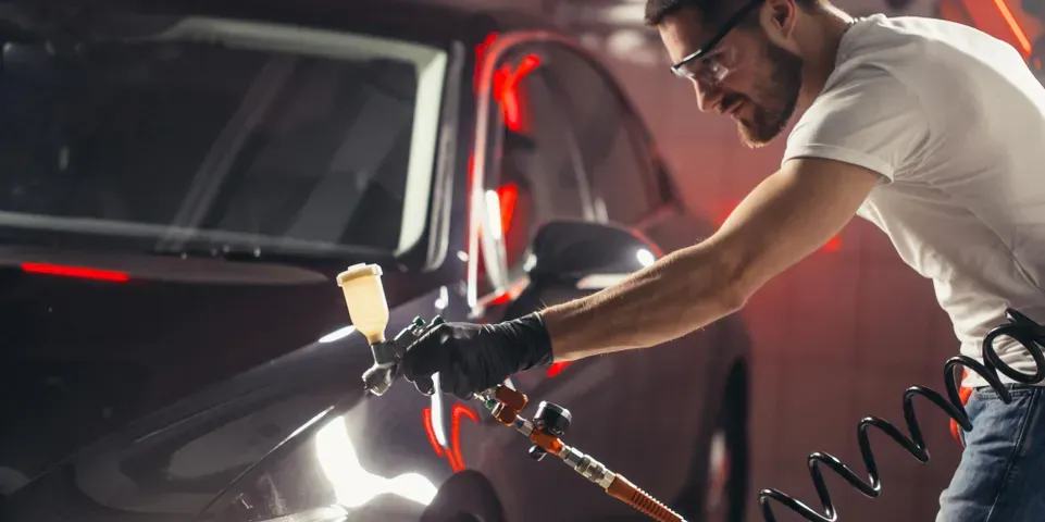 Man in a white shirt and glasses spraying a dark car, wearing black gloves in a garage with red lights.