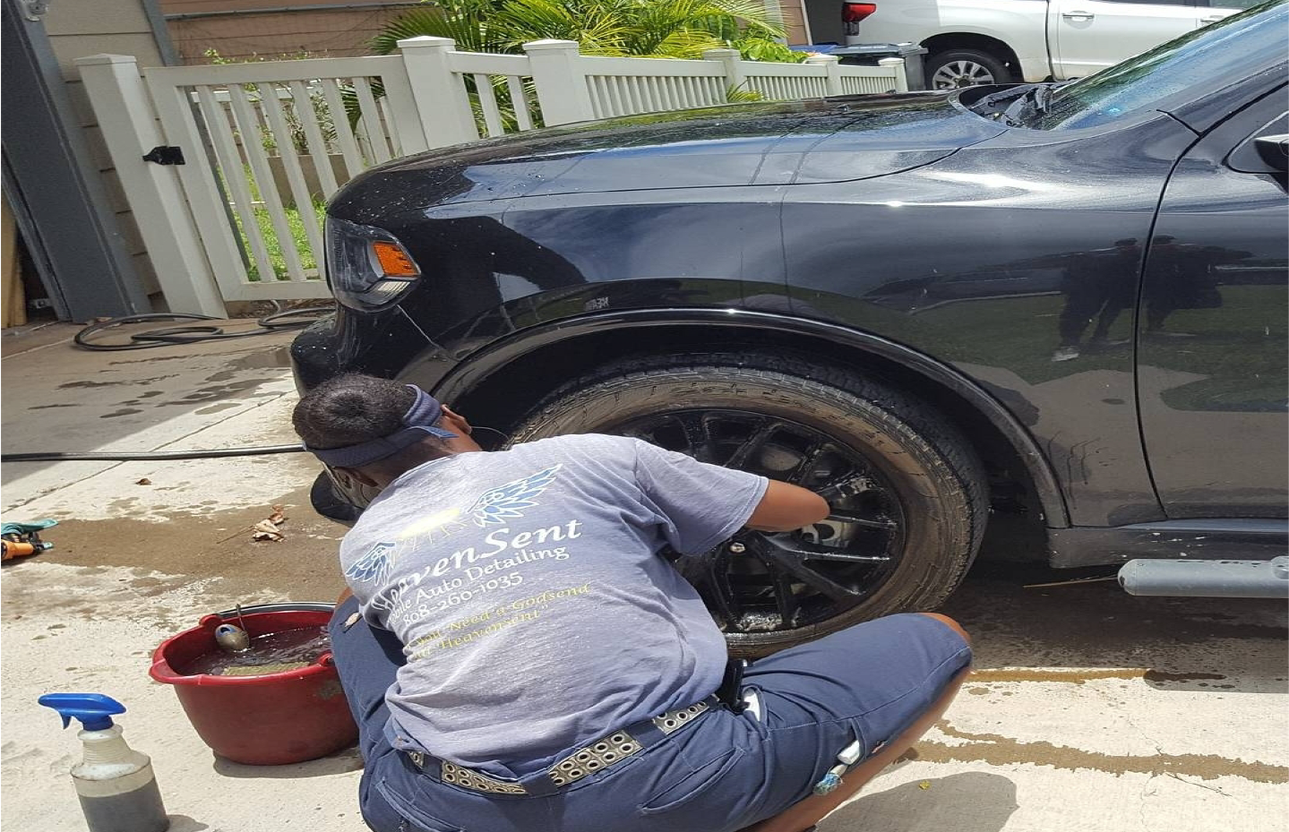 Person washing a black SUV's tire. They are kneeling, using a brush and bucket near a house and fence.