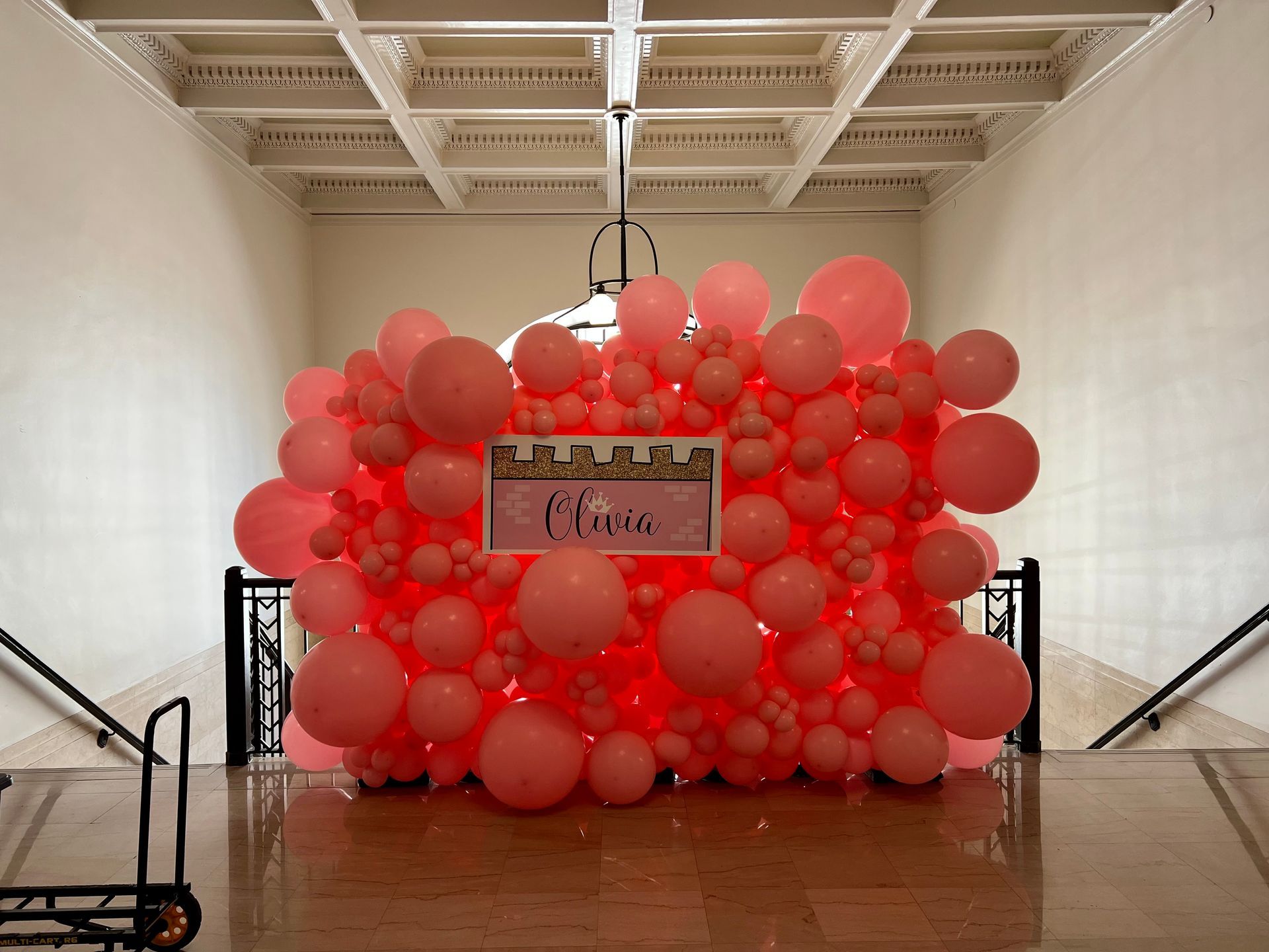 A wall of pink balloons in a room with stairs.