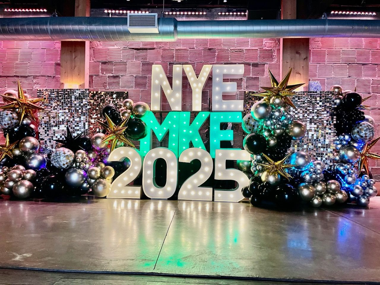 A large sign that says `` nye mke 2025 '' is surrounded by balloons and lights.