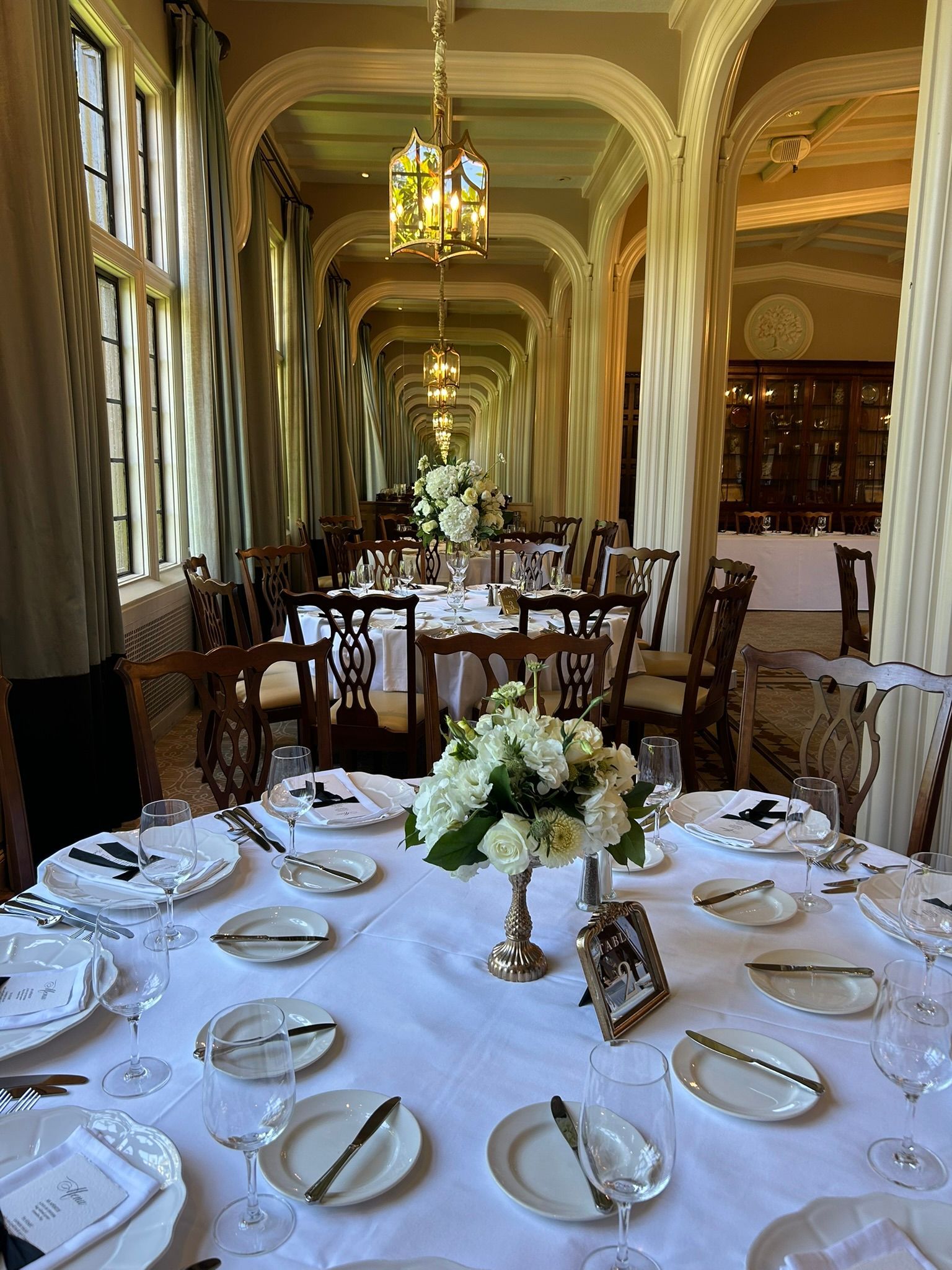 Formal dining room, round tables set with white linens, flowers, and place settings, long hall with tall windows.