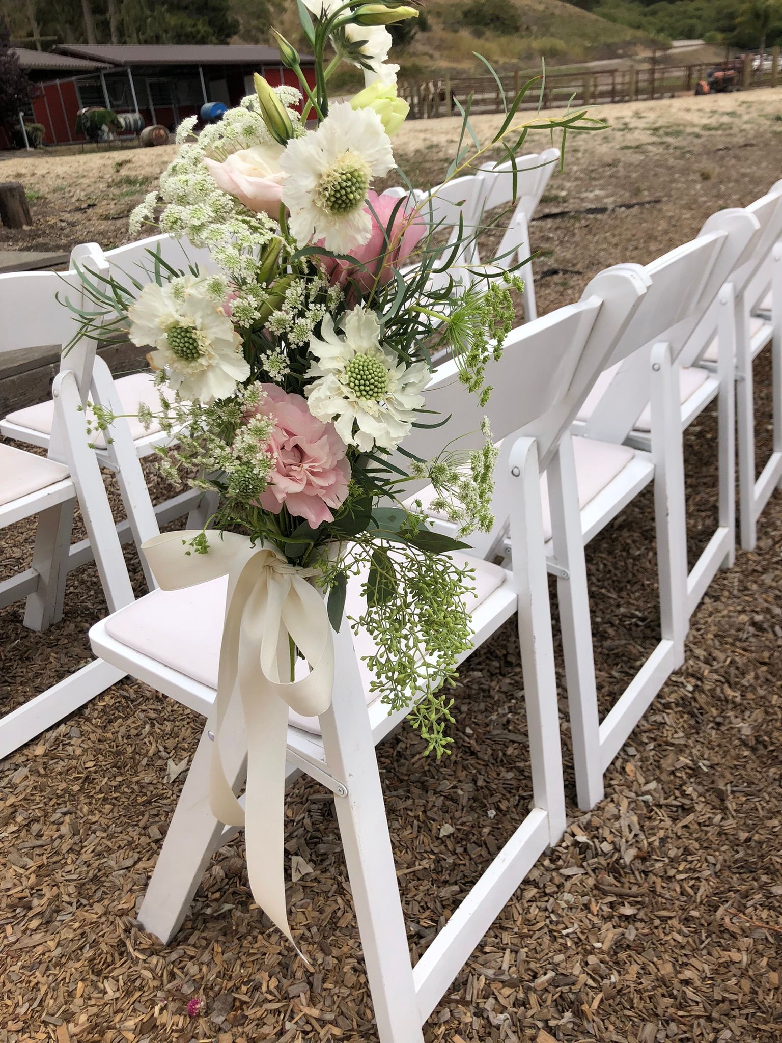 White folding chairs with floral arrangements tied to them, outdoor setting.