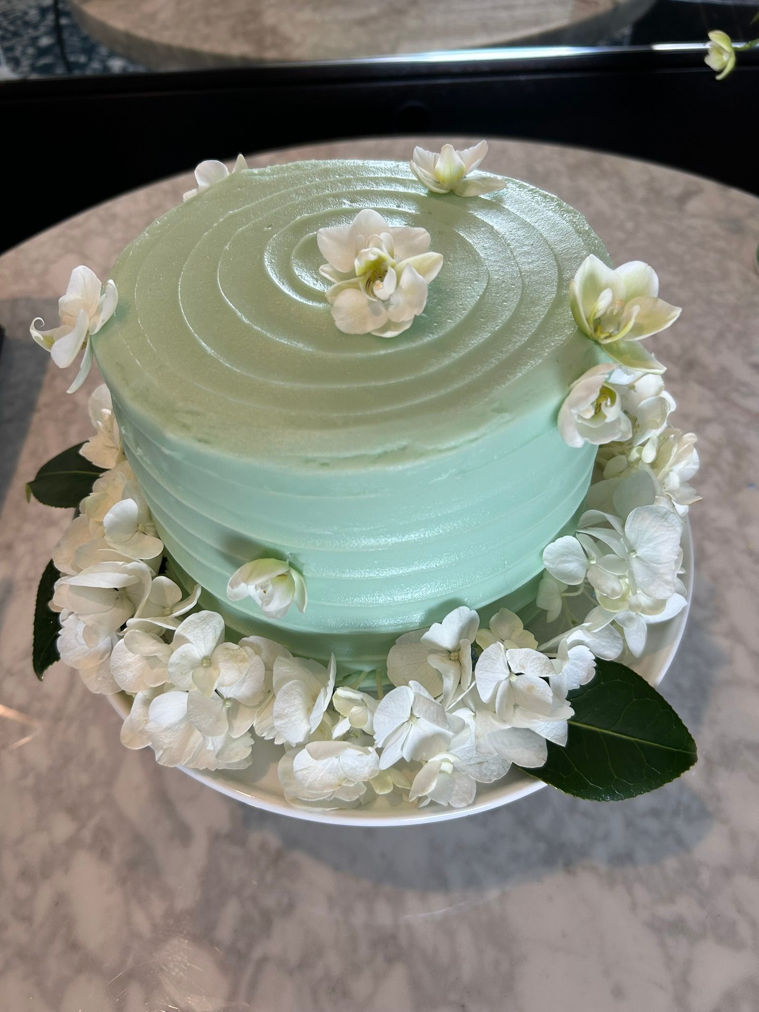 A mint green cake decorated with white flowers on a white plate, placed on a marble table.