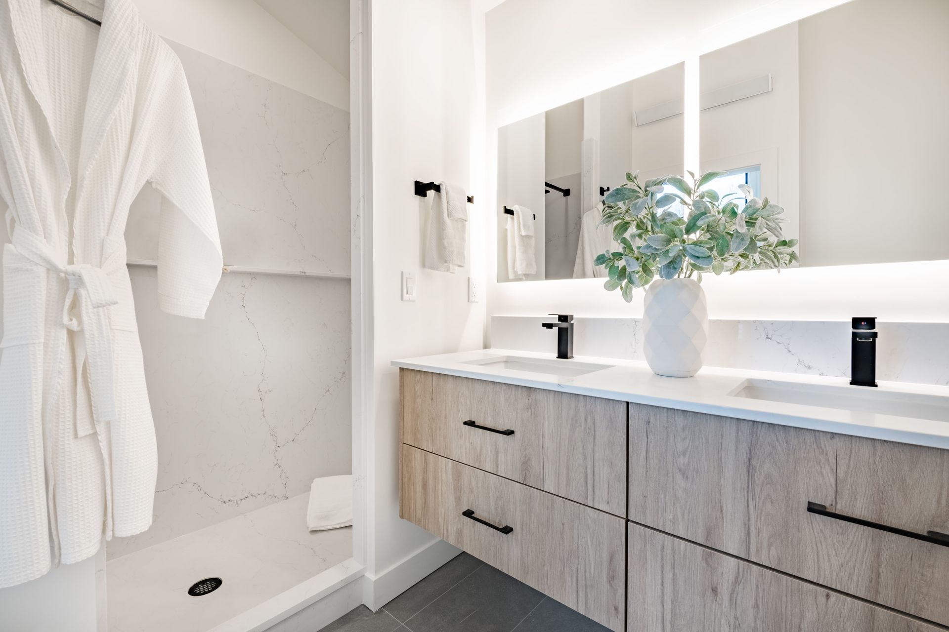 Bathroom with light wood vanity, white marble shower, robe, and a vase of greenery.