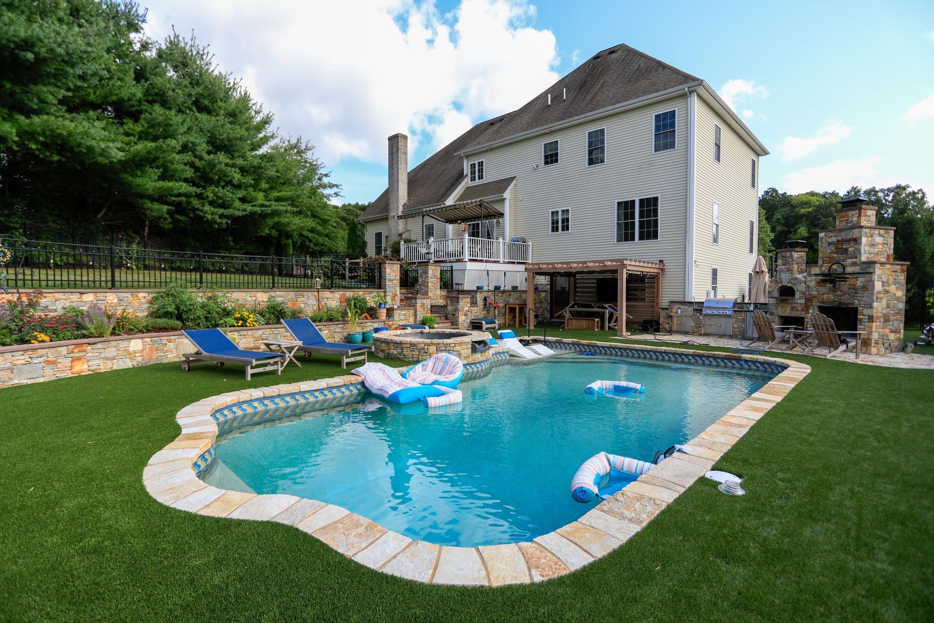 A backyard pool with floating inflatables, lounge chairs, a stone fireplace, and a large multi-story house in the background.