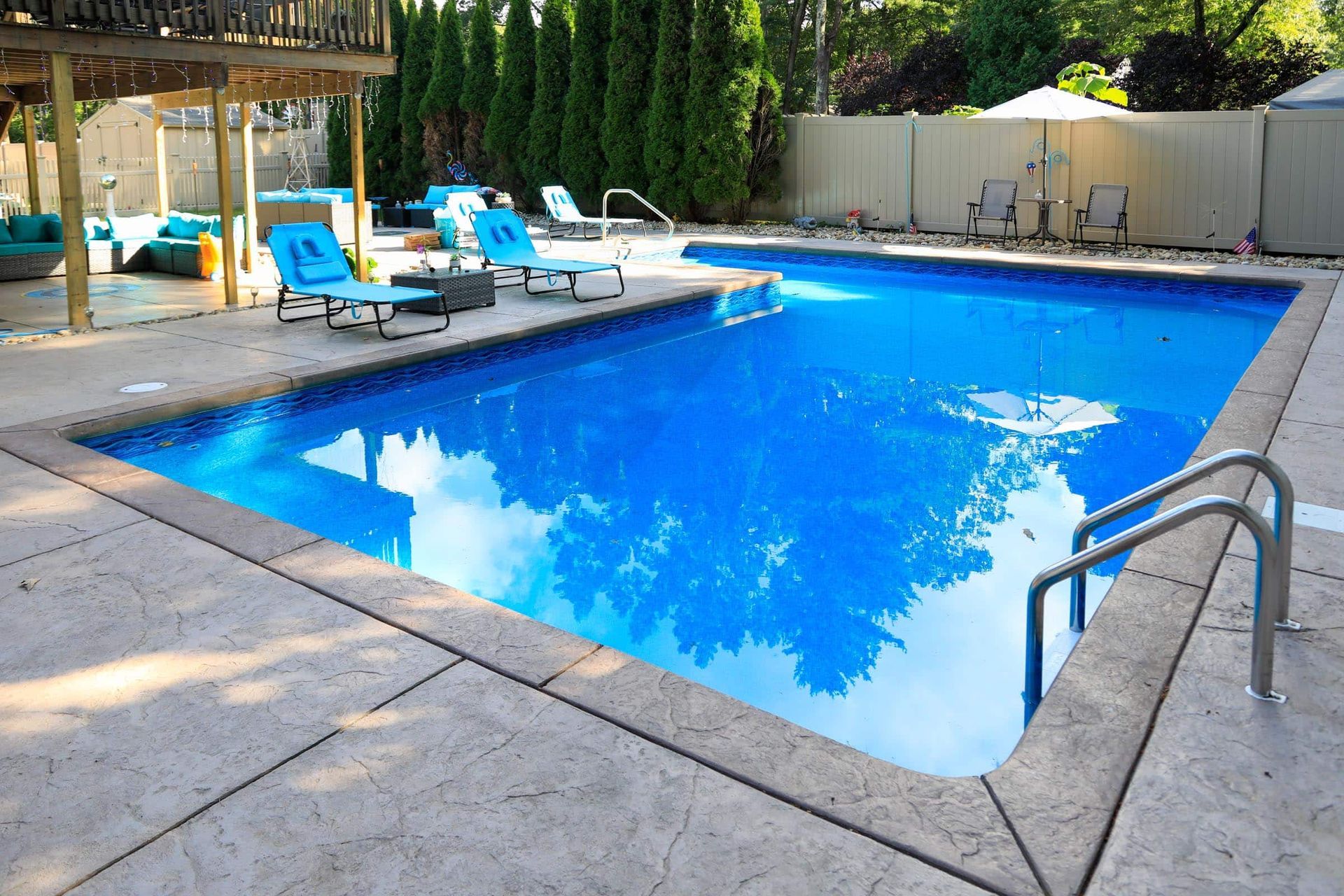 A bright blue rectangular backyard swimming pool surrounded by a concrete patio with lounge chairs and a wooden deck.