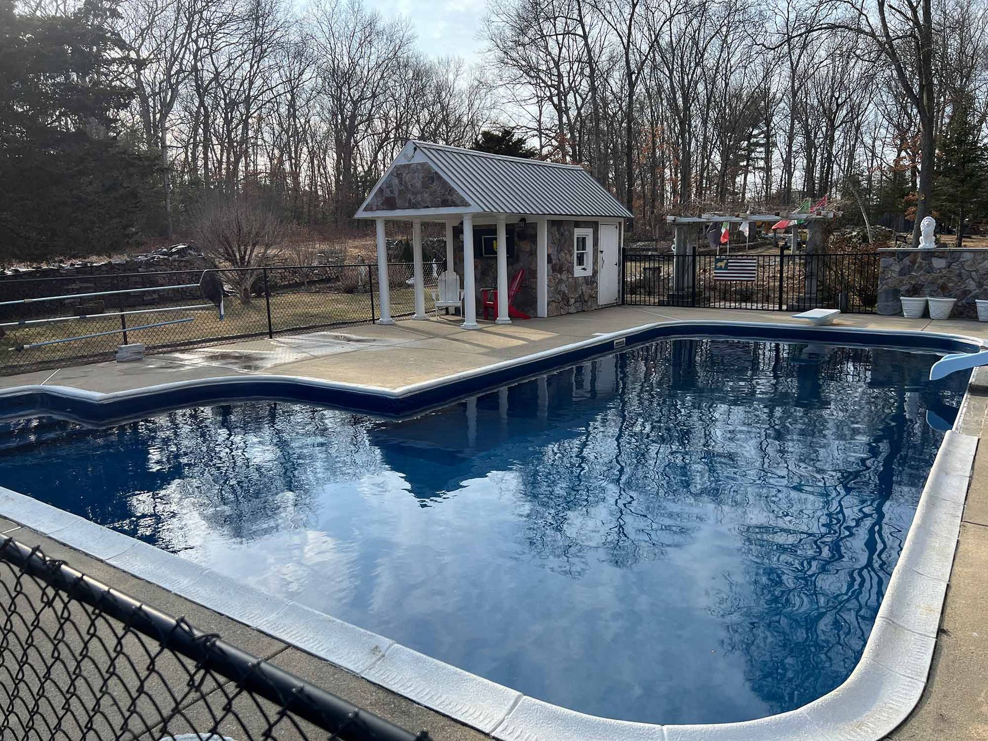 An outdoor rectangular swimming pool with a concrete deck, a small stone pool house, and surrounding trees.