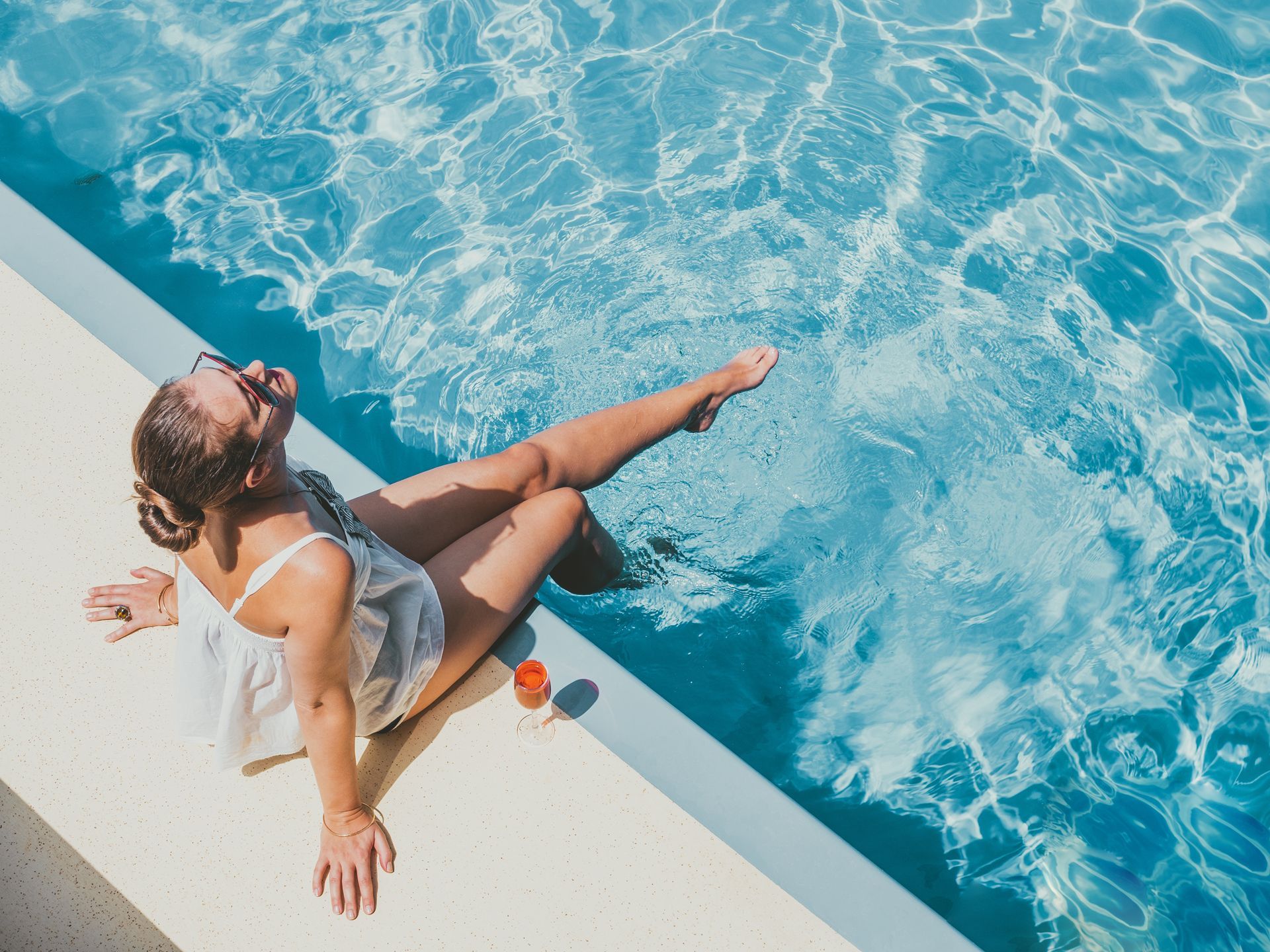 A person wearing a white top relaxes on the edge of a bright blue swimming pool, dipping one foot into the water.