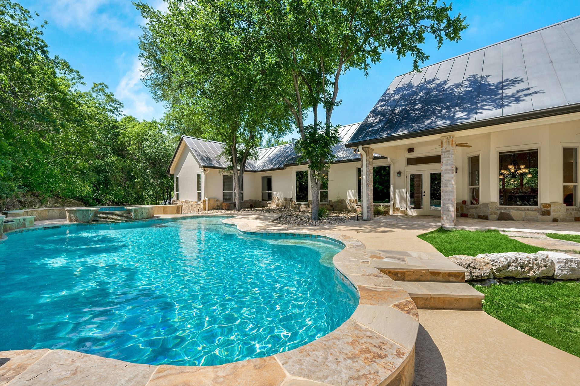A swimming pool with turquoise water sits next to a light-colored, multi-roofed house surrounded by trees.