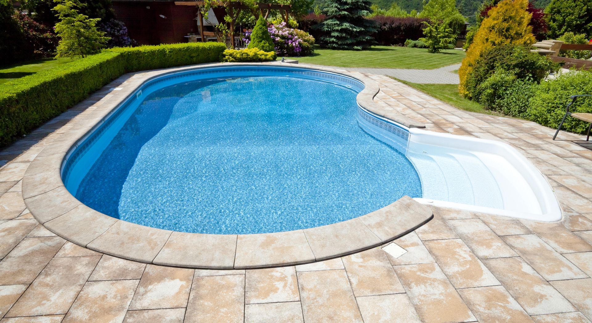 A blue, kidney-shaped swimming pool with a white stepped entry in a stone-paved backyard with a manicured hedge.