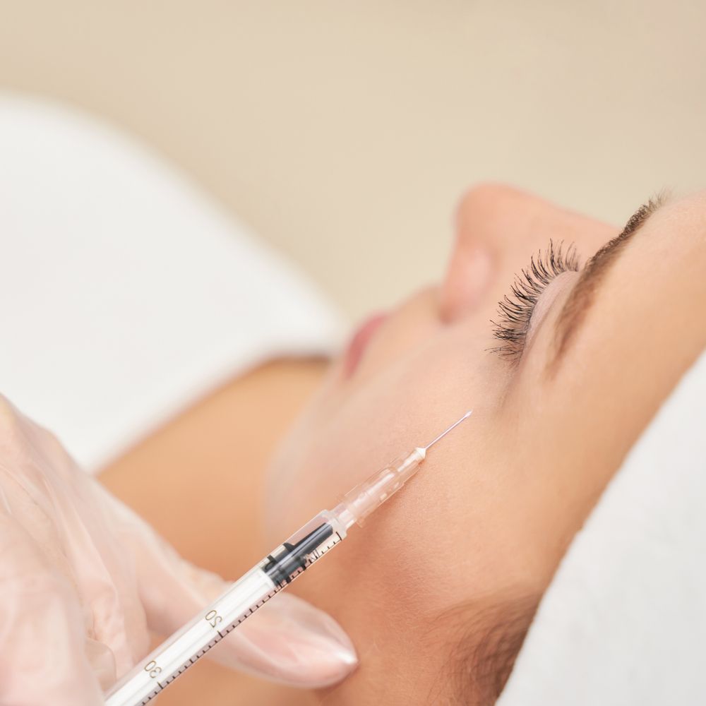 A gloved hand injects a syringe near a person's cheek during a cosmetic procedure.