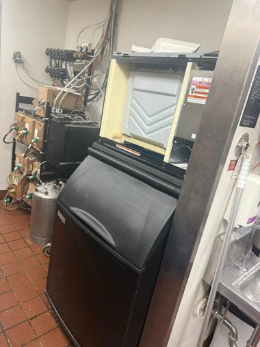 Commercial kitchen: ice machine, equipment, a metal sink, and various pipes against a wall.