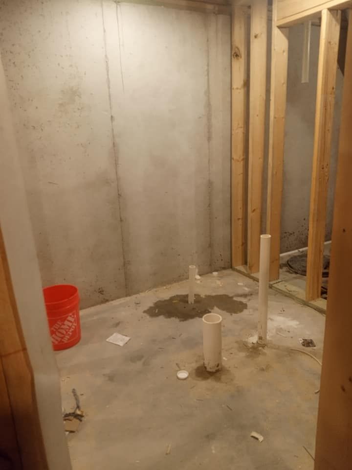 Unfinished basement with exposed concrete walls, framing, and plumbing.  A bucket sits on the floor.