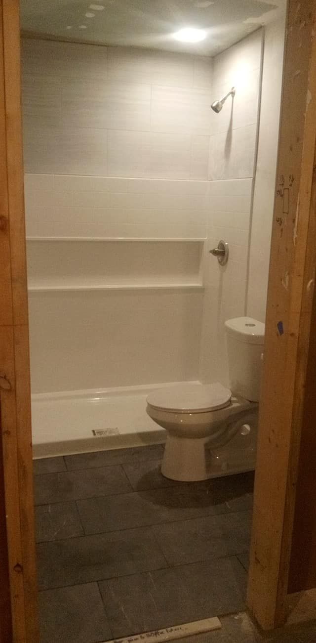A small bathroom with a white shower, toilet, and gray flooring. The wooden door frame is visible.