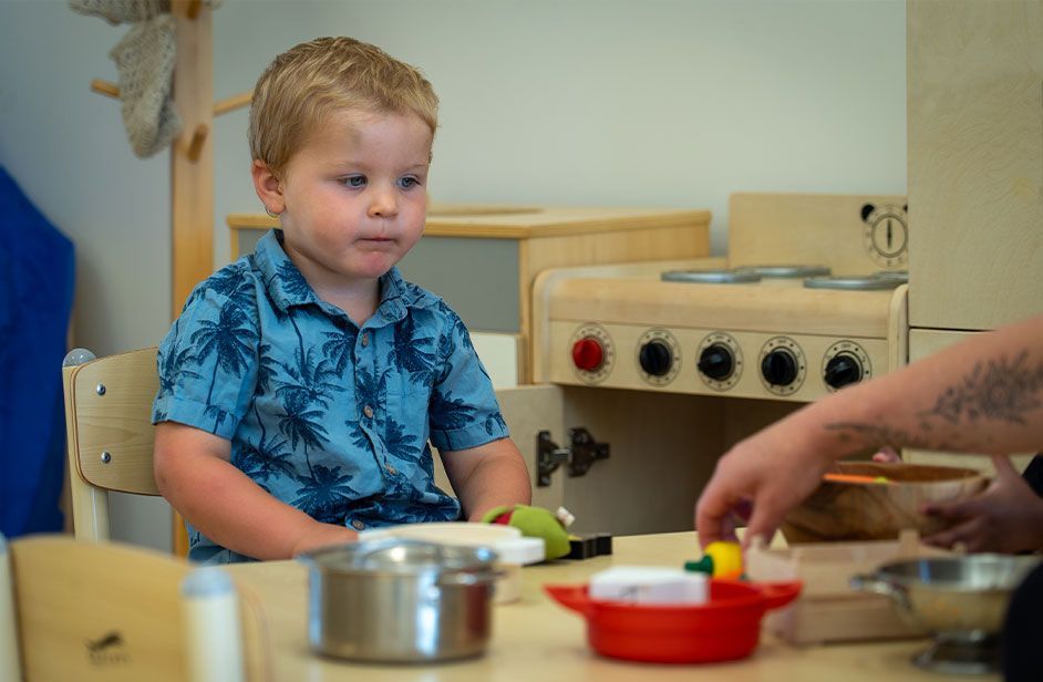Toddler and carer in play kitchen | Carer demonstating play | Toddler watching carers hand intently