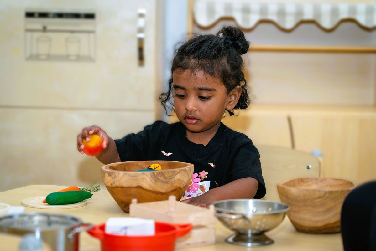 Toddler in play kitchen playing with wooden fruit in right hand | Wooden mixing bowl on table