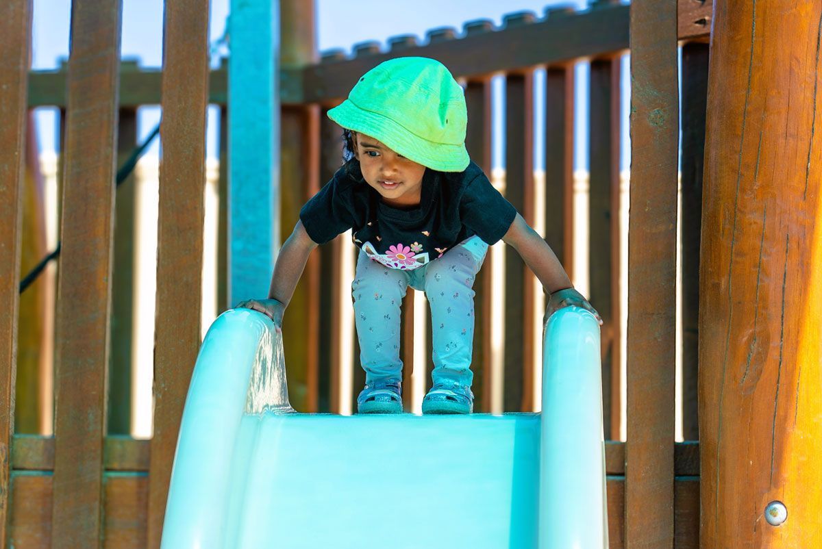Toddler in oversized green hat at top of blue slide | Toddler playing outside