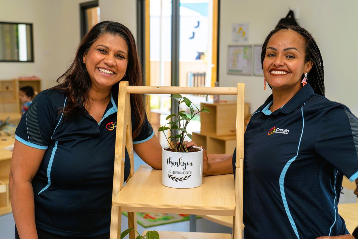 Two Carers linking arms behind pot plant on wooden stand | Writing on pot | thankyou for helping me grow
