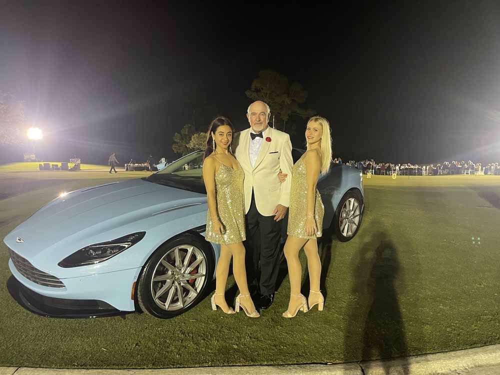 A man and two women are standing in front of a blue car.