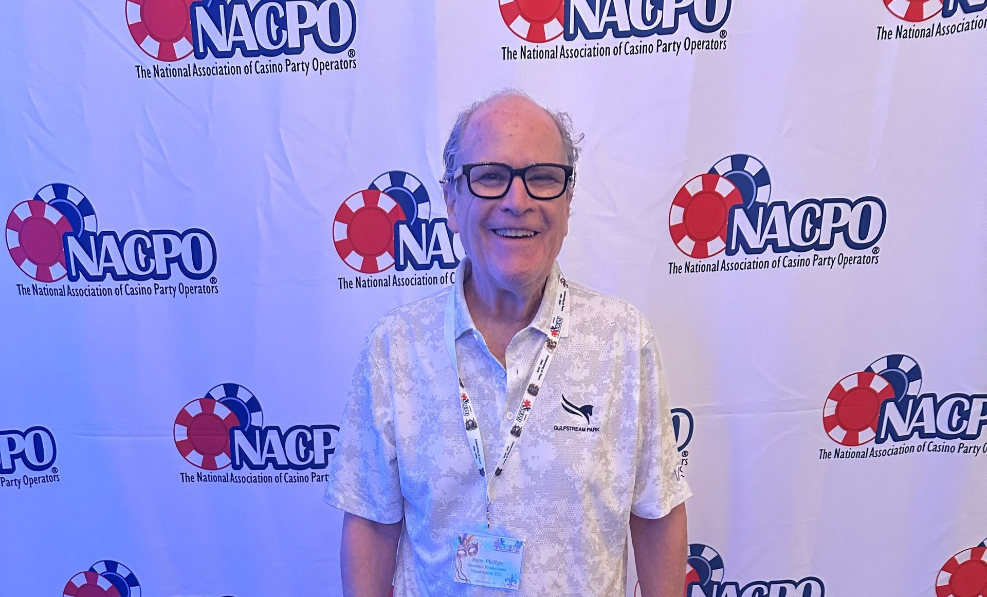 Man standing and smiling in front of a branded NAPO backdrop, wearing a floral shirt, shorts, and sneakers
