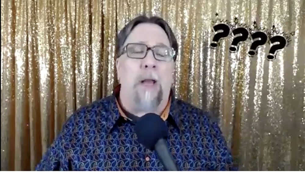 A man with glasses and a beard is talking into a microphone in front of a gold curtain.