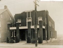 Sepia-toned photo of a two-story brick building on a street corner with utility poles and sidewalks.