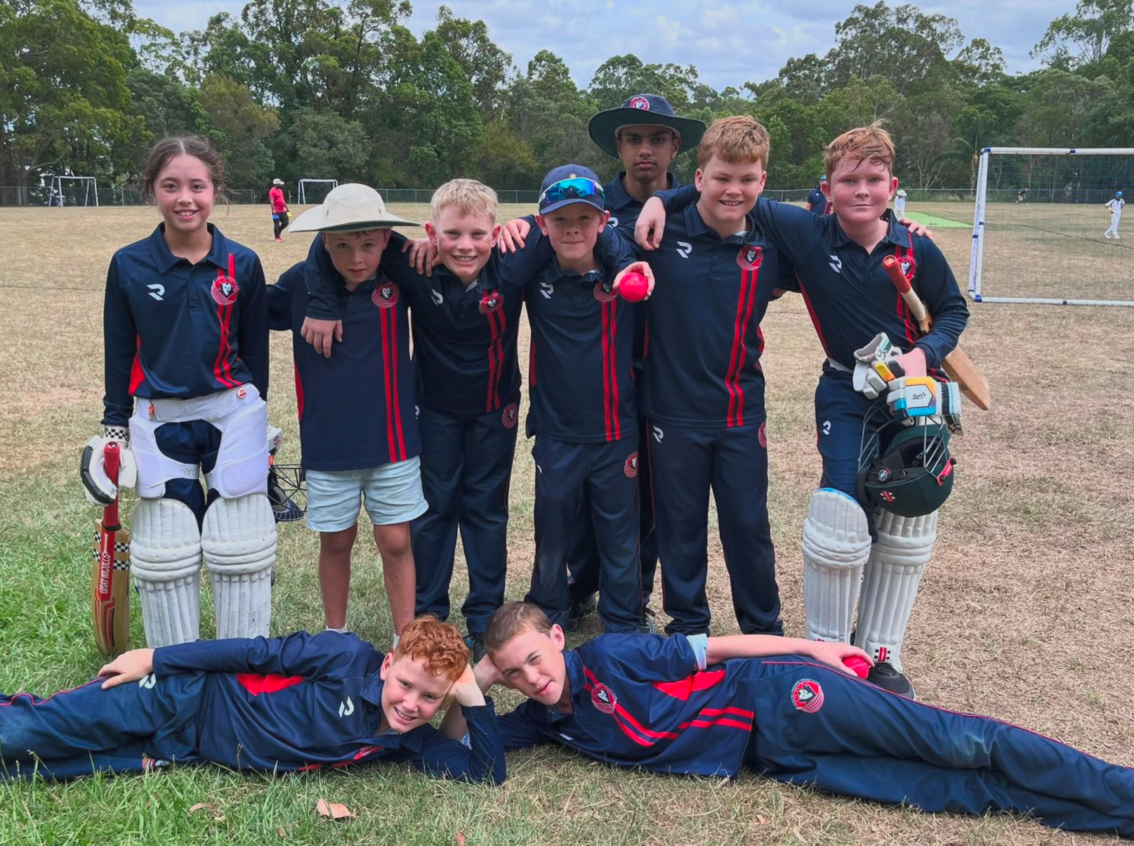 Cricket team posing on a field, with two players lying in front and wickets visible behind them.