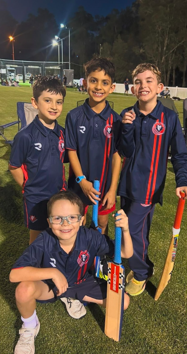 Four children in matching sports jerseys pose on a grassy field at night with cricket bats and stumps.