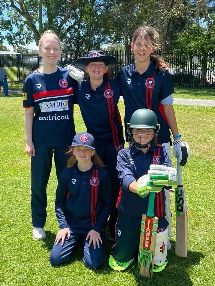 Five cricket players in navy jerseys posing on a grassy field with bats and gear under trees