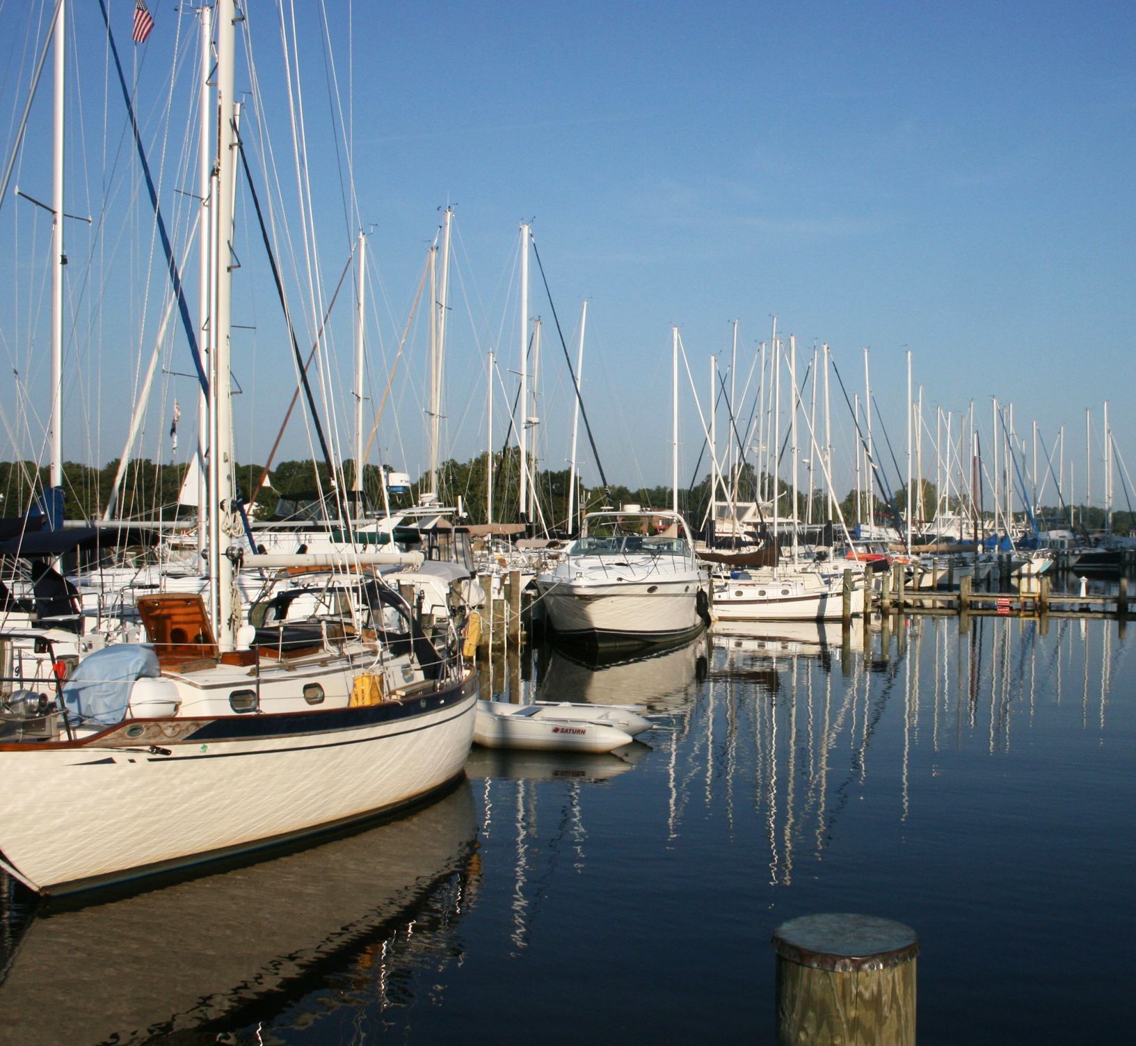 Sailboats docked in a marina, masts reflected in calm water under a blue sky.