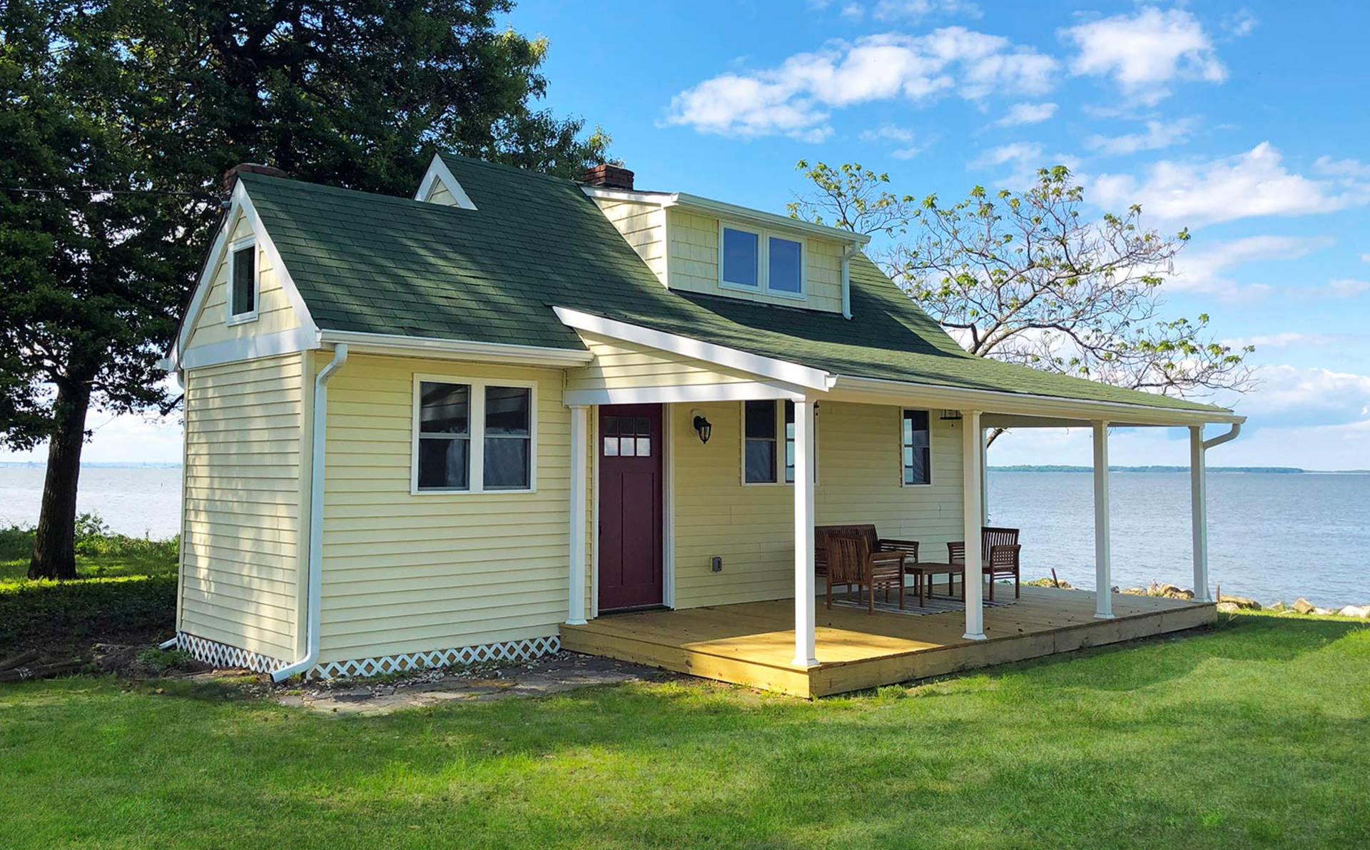 Yellow house with green roof and porch overlooking a body of water.