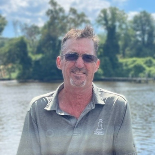 Man wearing sunglasses and a gray polo shirt, smiling by water with trees in the background.