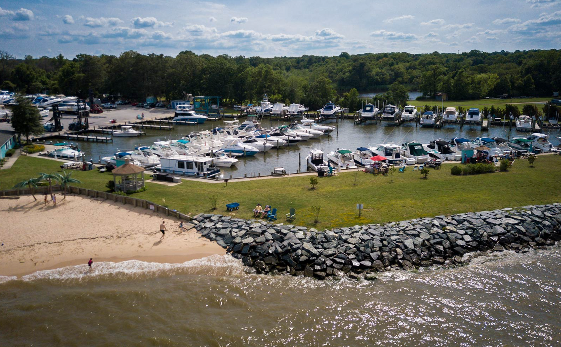 Boats docked at a marina next to a small beach. People on the beach and green grass area. Blue sky with clouds.
