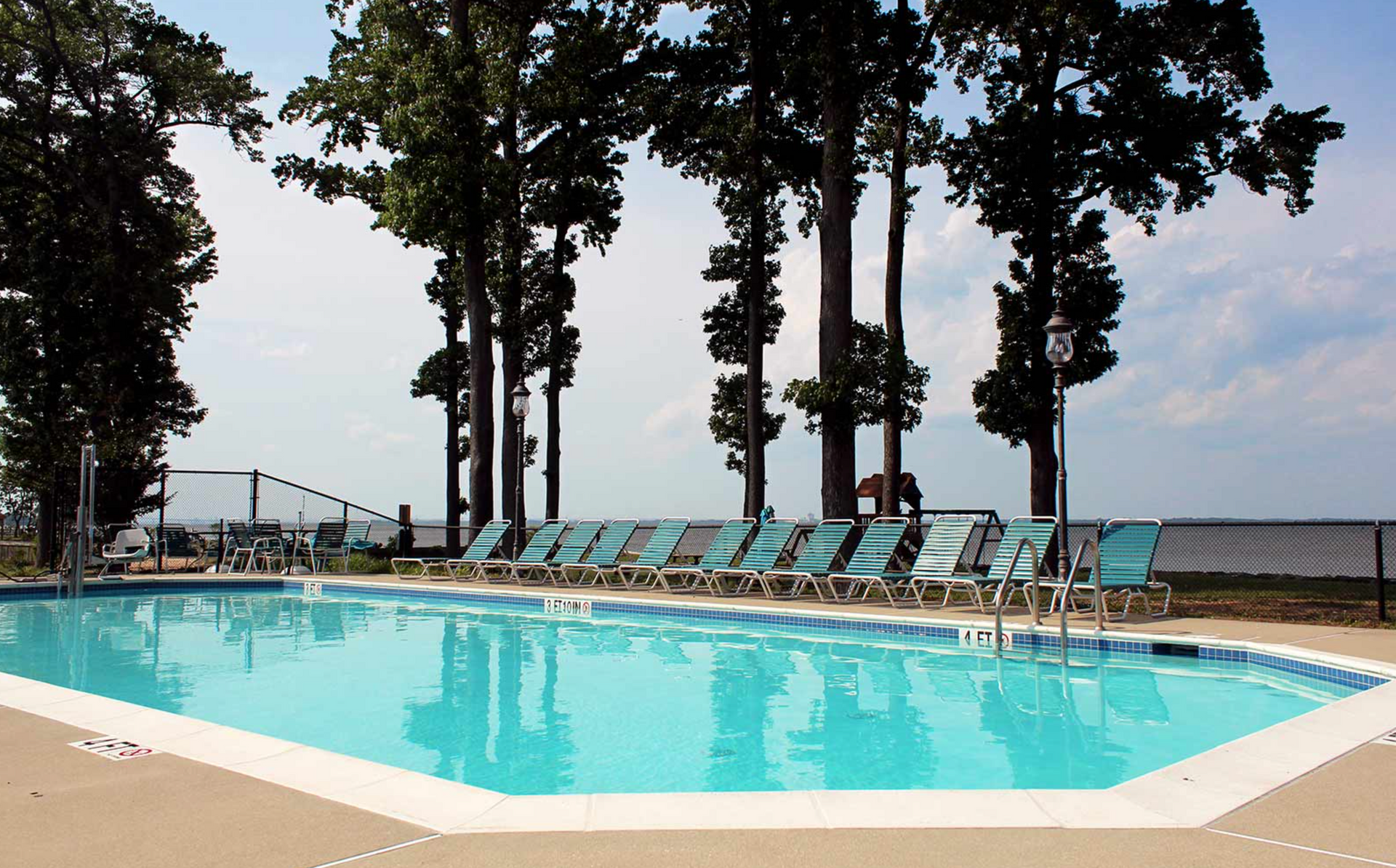 Swimming pool with lounge chairs, tall trees, and a fence.