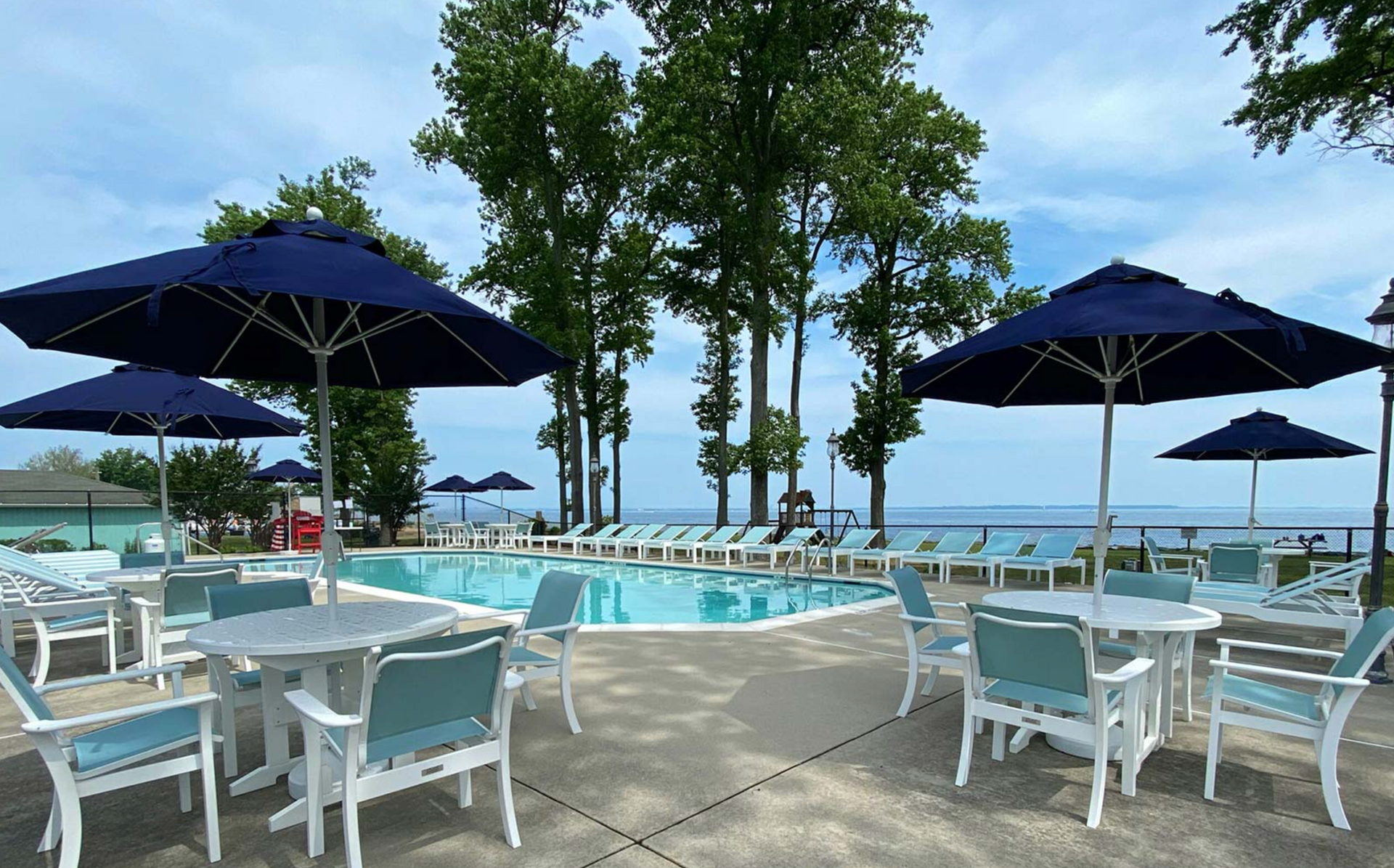 Poolside scene with tables, chairs, and umbrellas. Trees and water in the background under a cloudy sky.