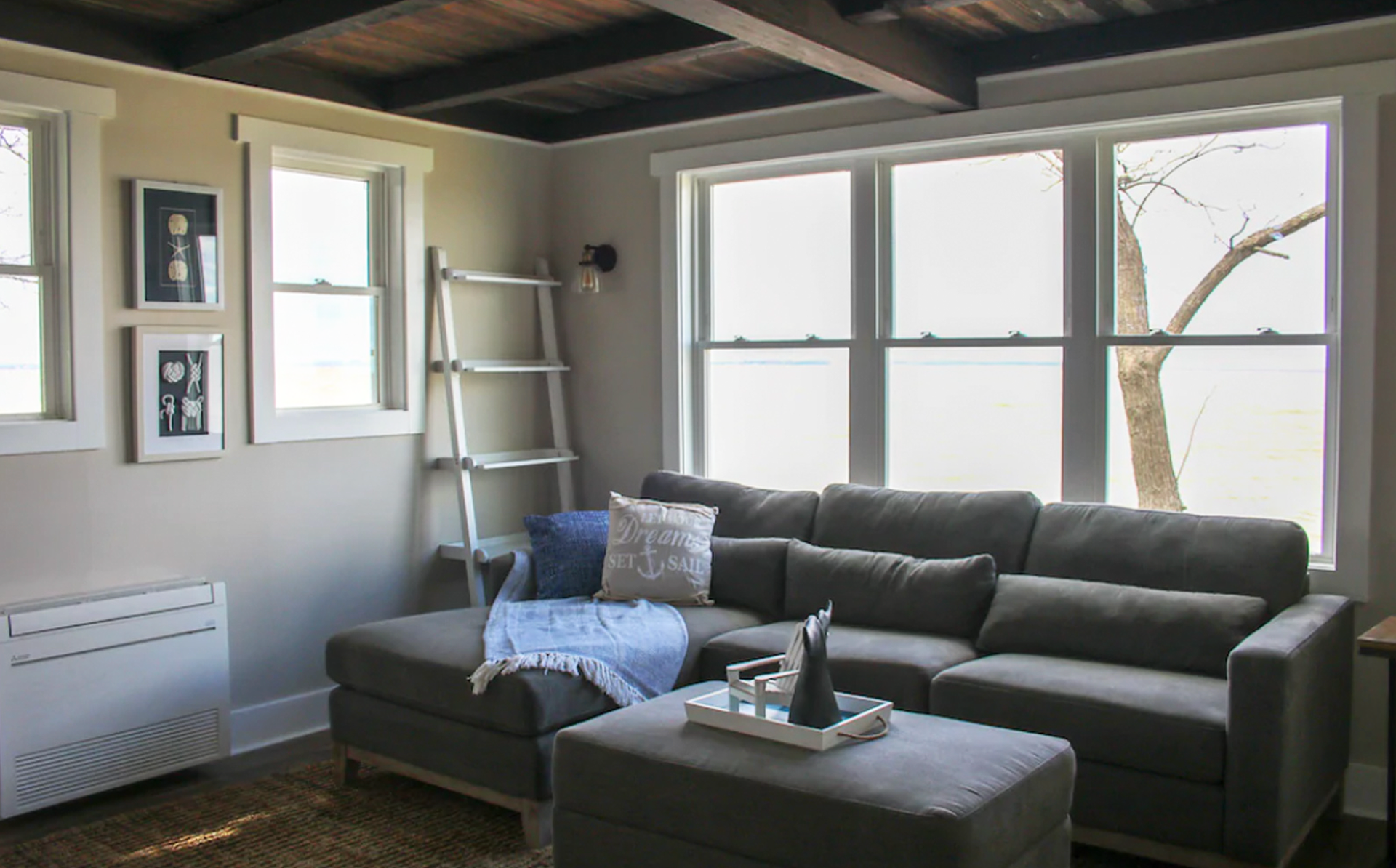 Cozy living room with gray sectional sofa, ottoman, and large windows overlooking a tree.
