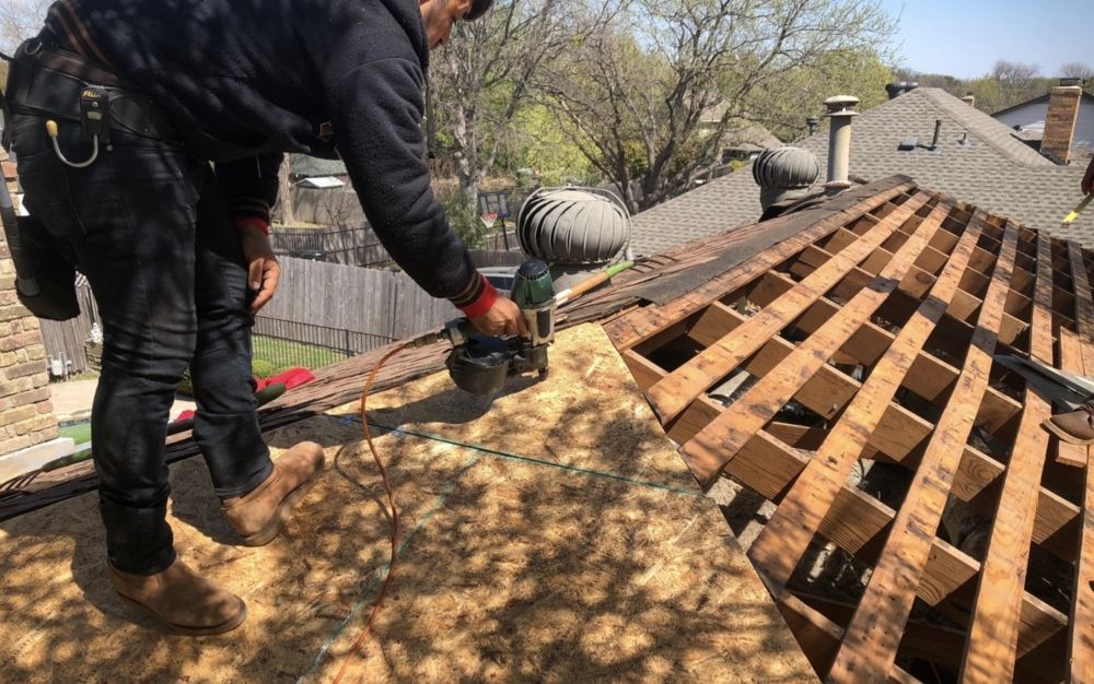 Workers removing roof shingles with a circular saw on a house roof, exposing wooden rafters.