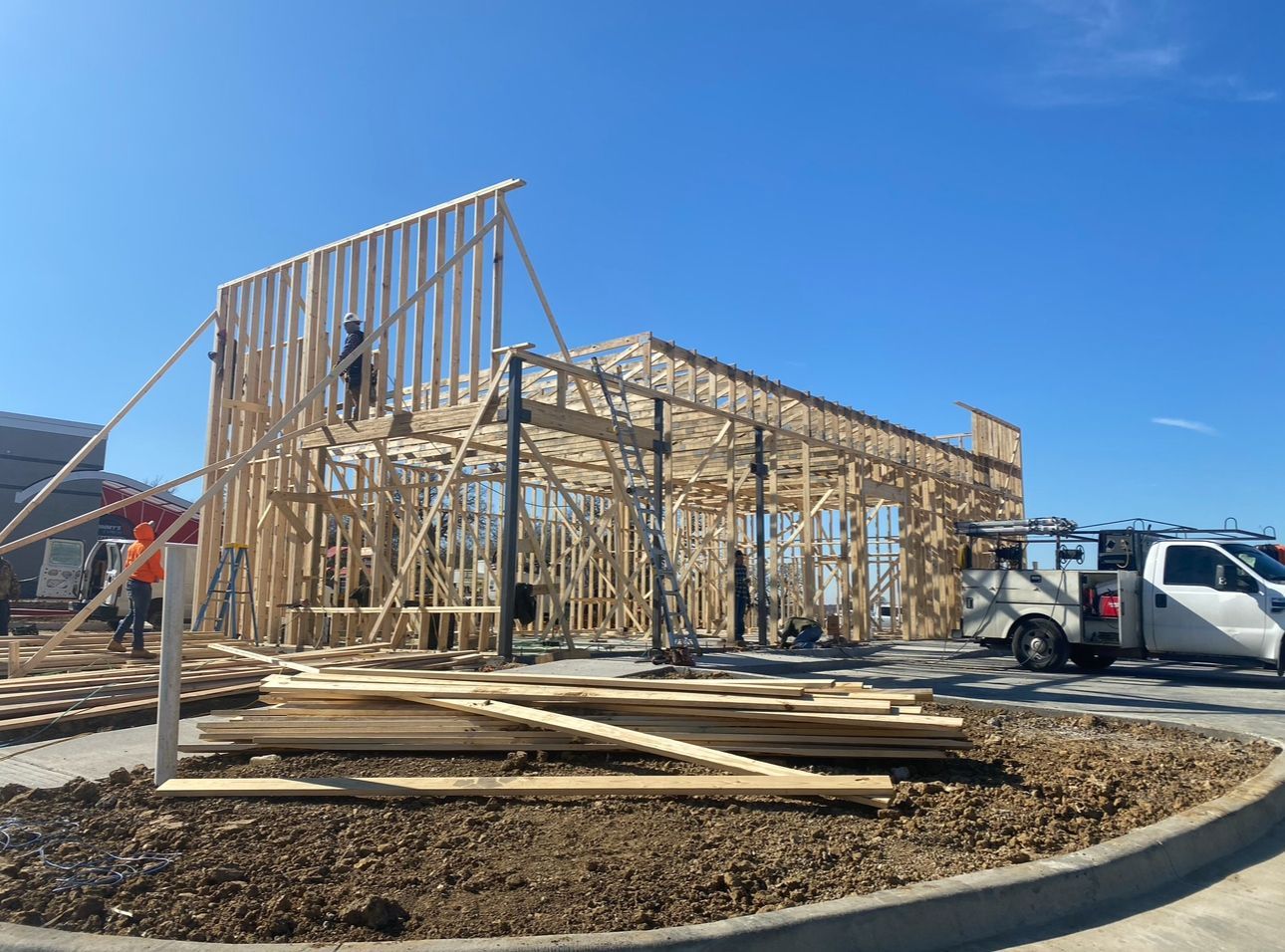Wood-frame house under construction on a sunny day, with a truck and lumber piles in the foreground.