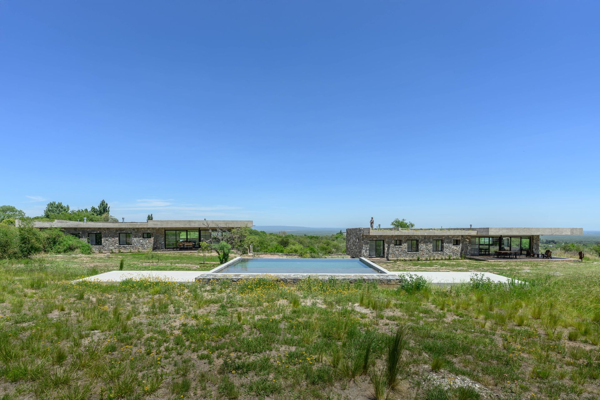 Dos edificios de piedra con piscina, situados en un paisaje verde bajo un cielo azul.