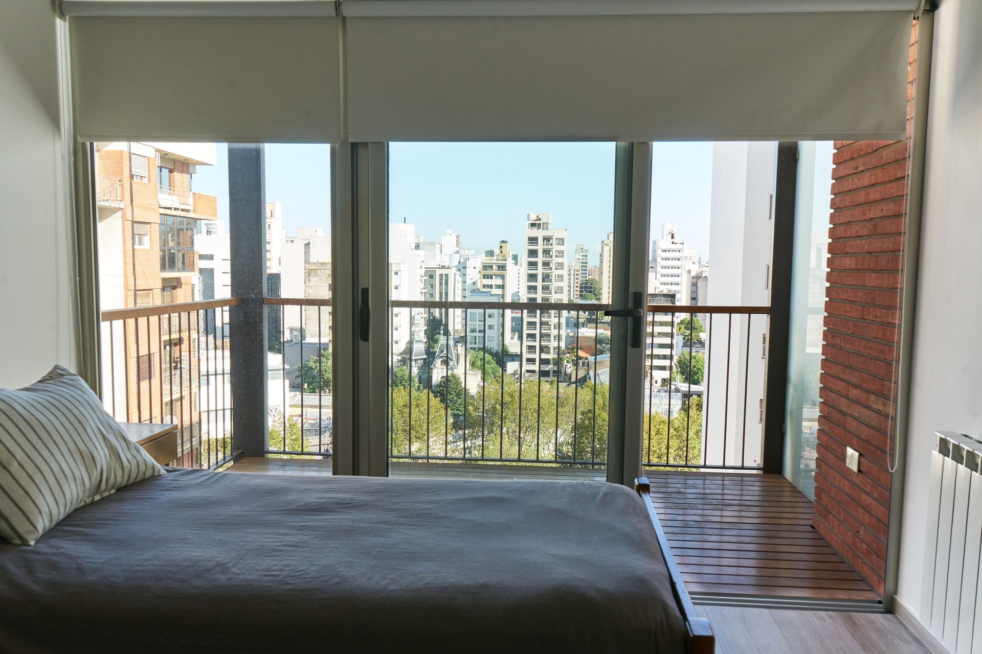 Dormitorio con cama frente a puertas abiertas con vista a la ciudad, cortinas enrollables color beige claro.