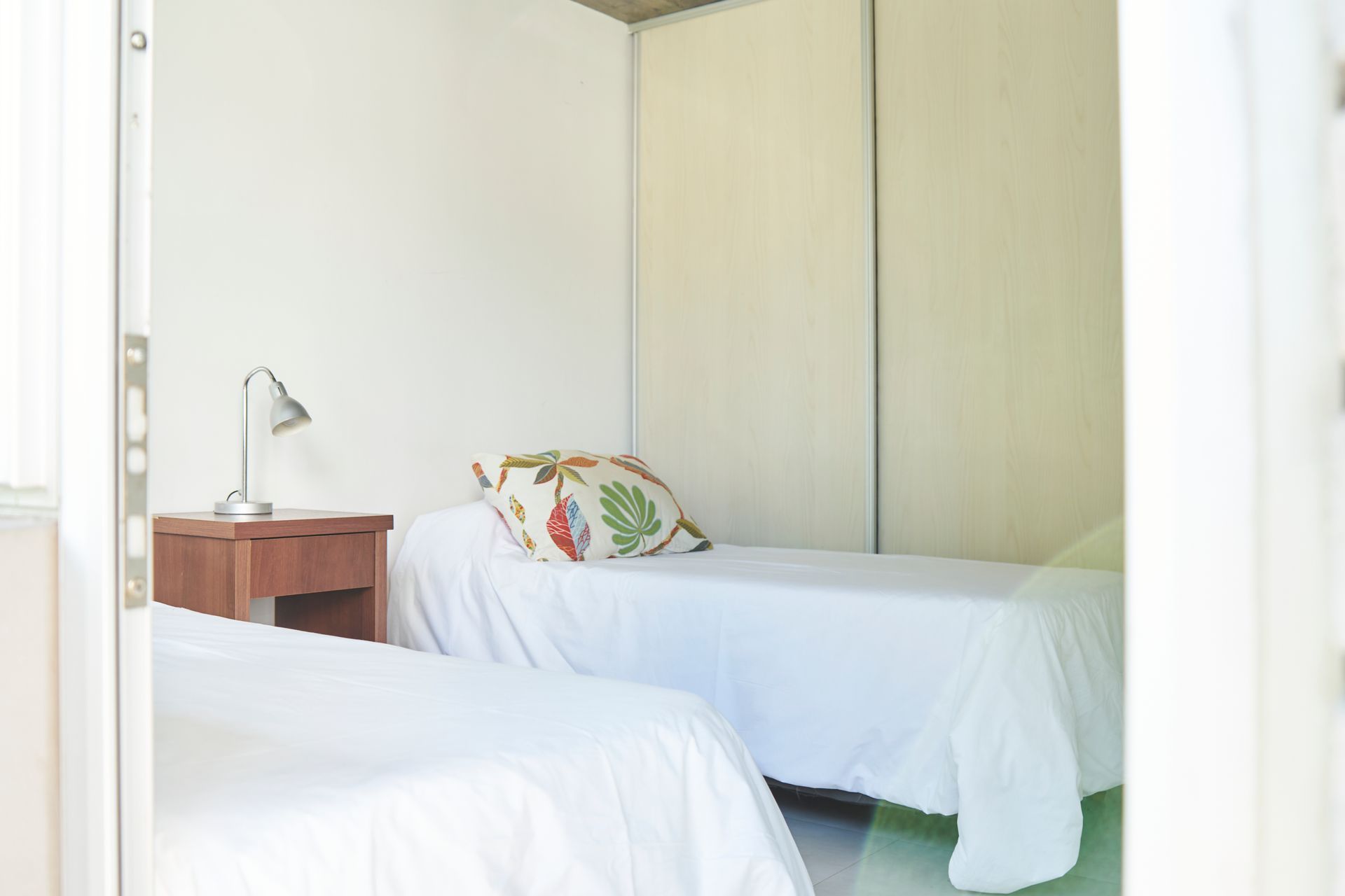 Dos camas individuales en una habitación sencilla; ropa de cama blanca, mesa de noche con lámpara, almohada y armario.