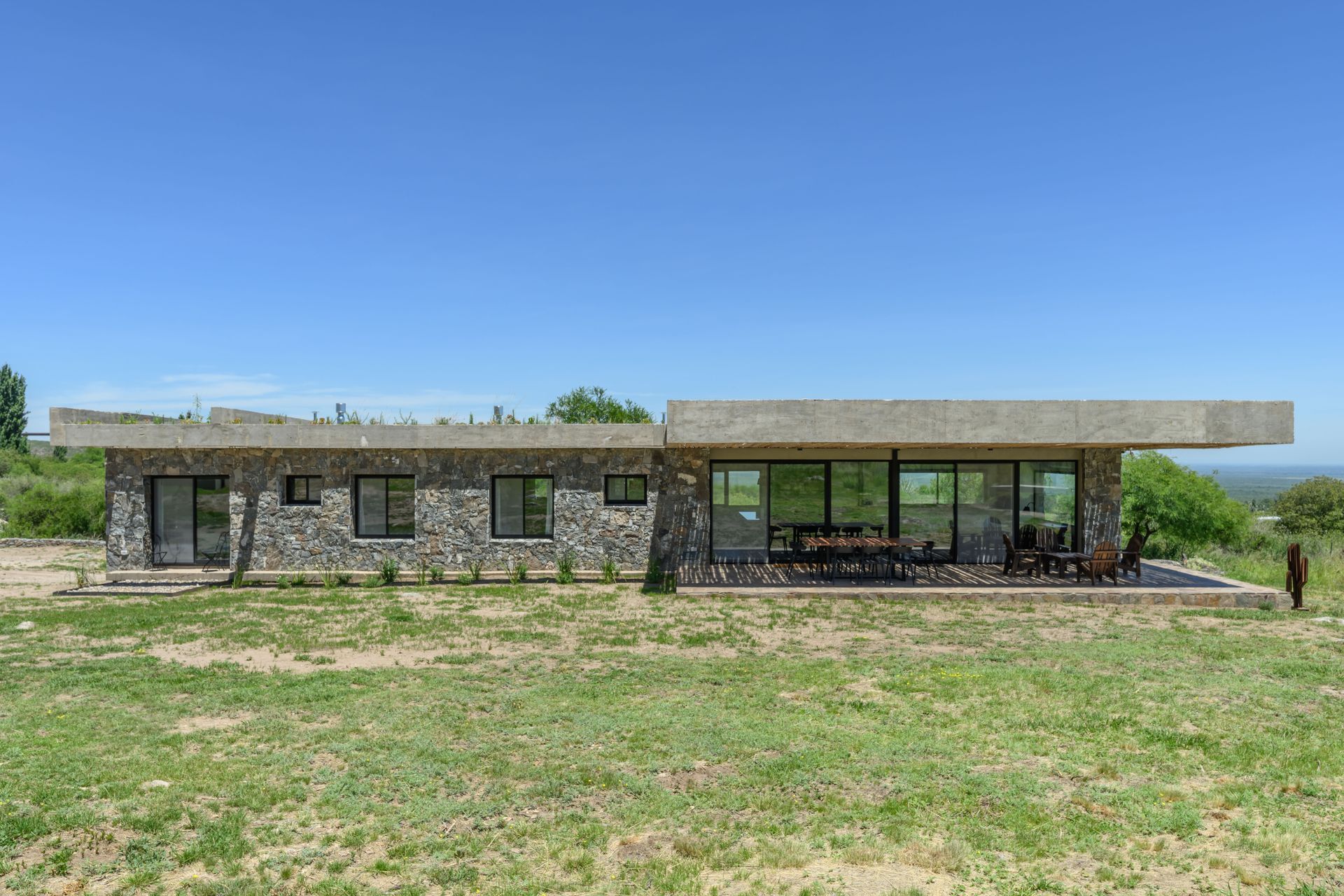 Casa de piedra con techo de hormigón, porche con mesa y sillas, situada en un campo de hierba bajo un cielo azul.