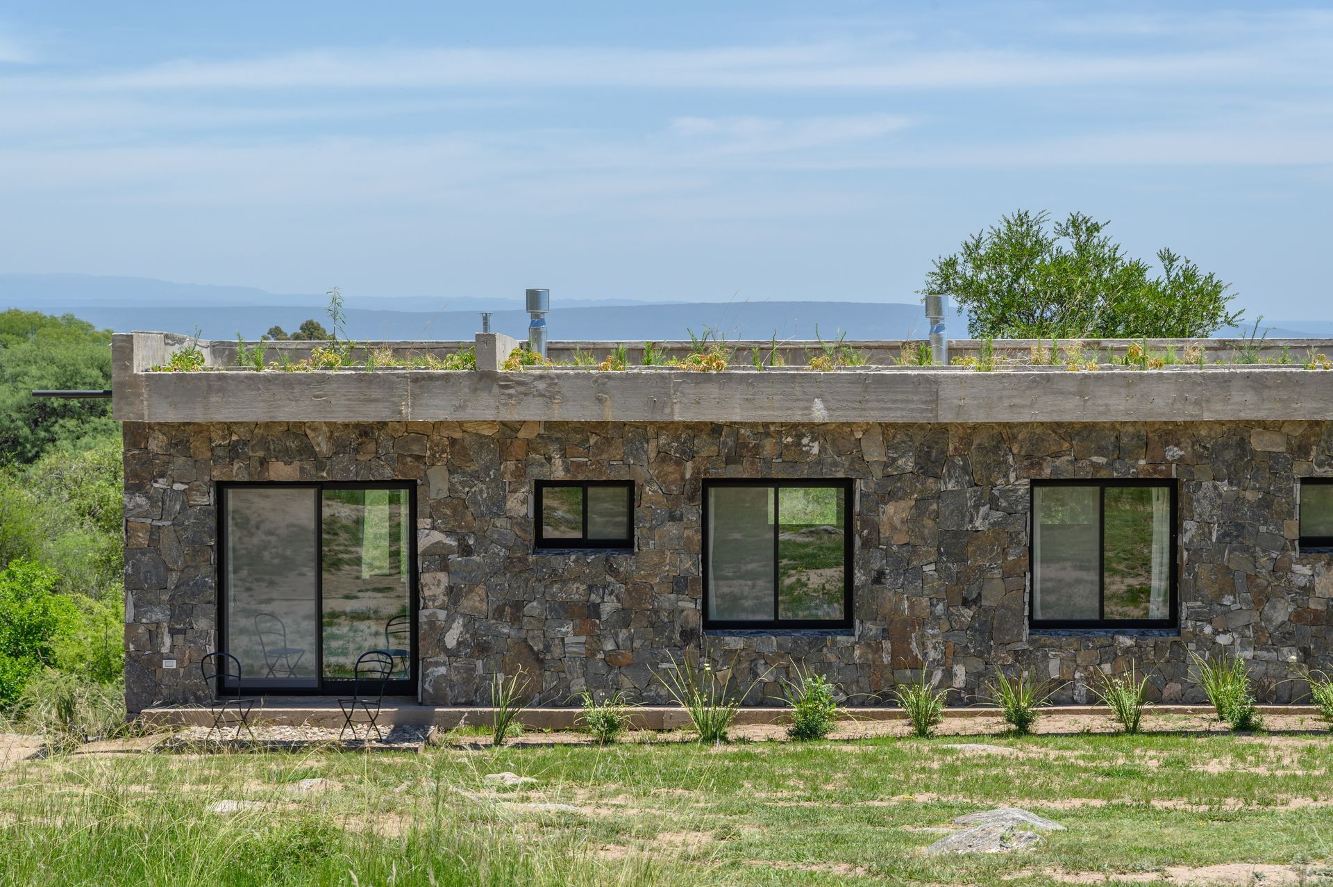 Casa con fachada de piedra, ventanas con marcos negros y techo plano con vegetación, enclavada en un paisaje natural.