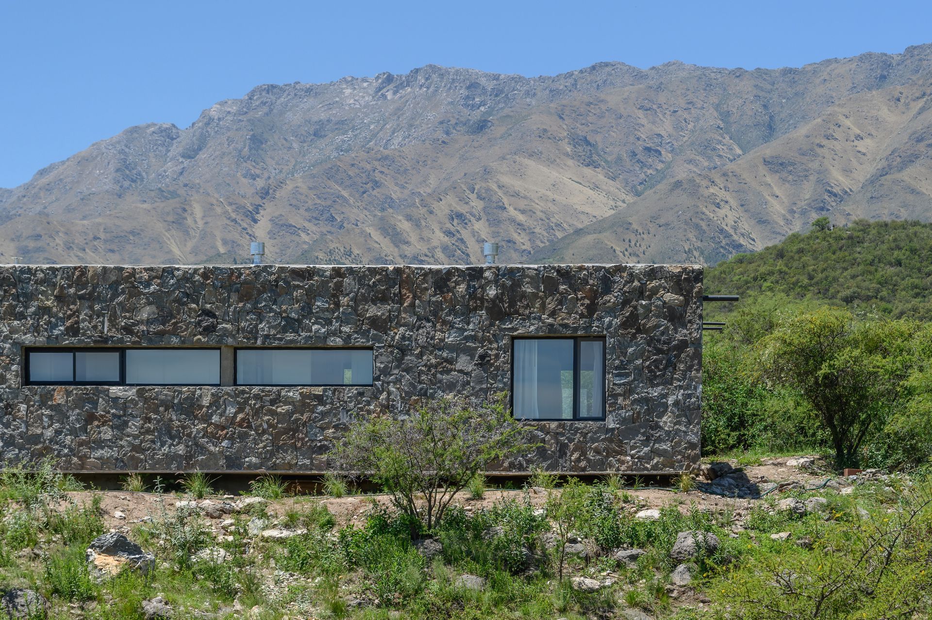Casa moderna revestida de piedra con ventanas rectangulares, situada en un paisaje montañoso.