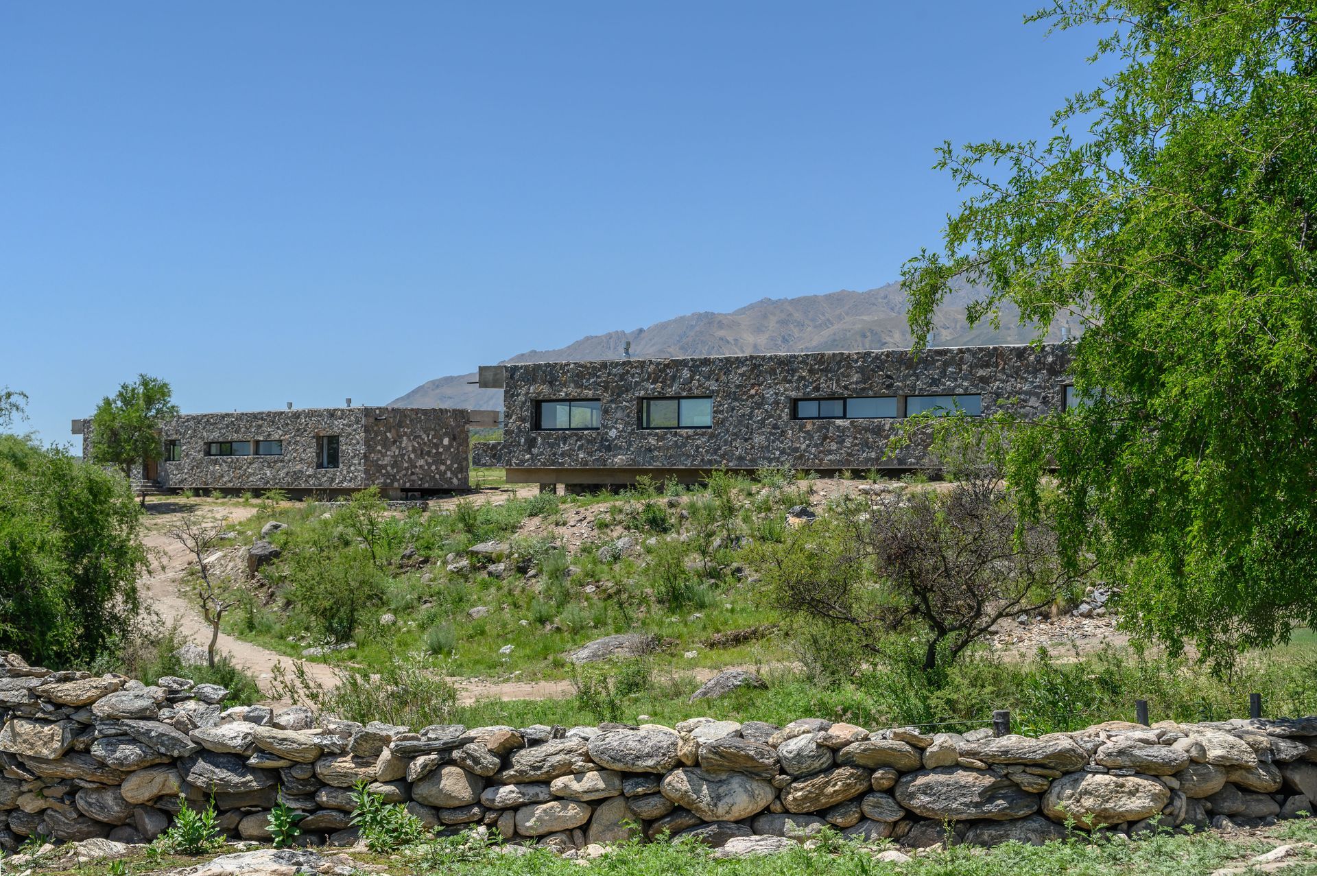 Edificios modernos con fachadas de piedra en una ladera bajo un cielo azul, parcialmente oscurecido por la vegetación.