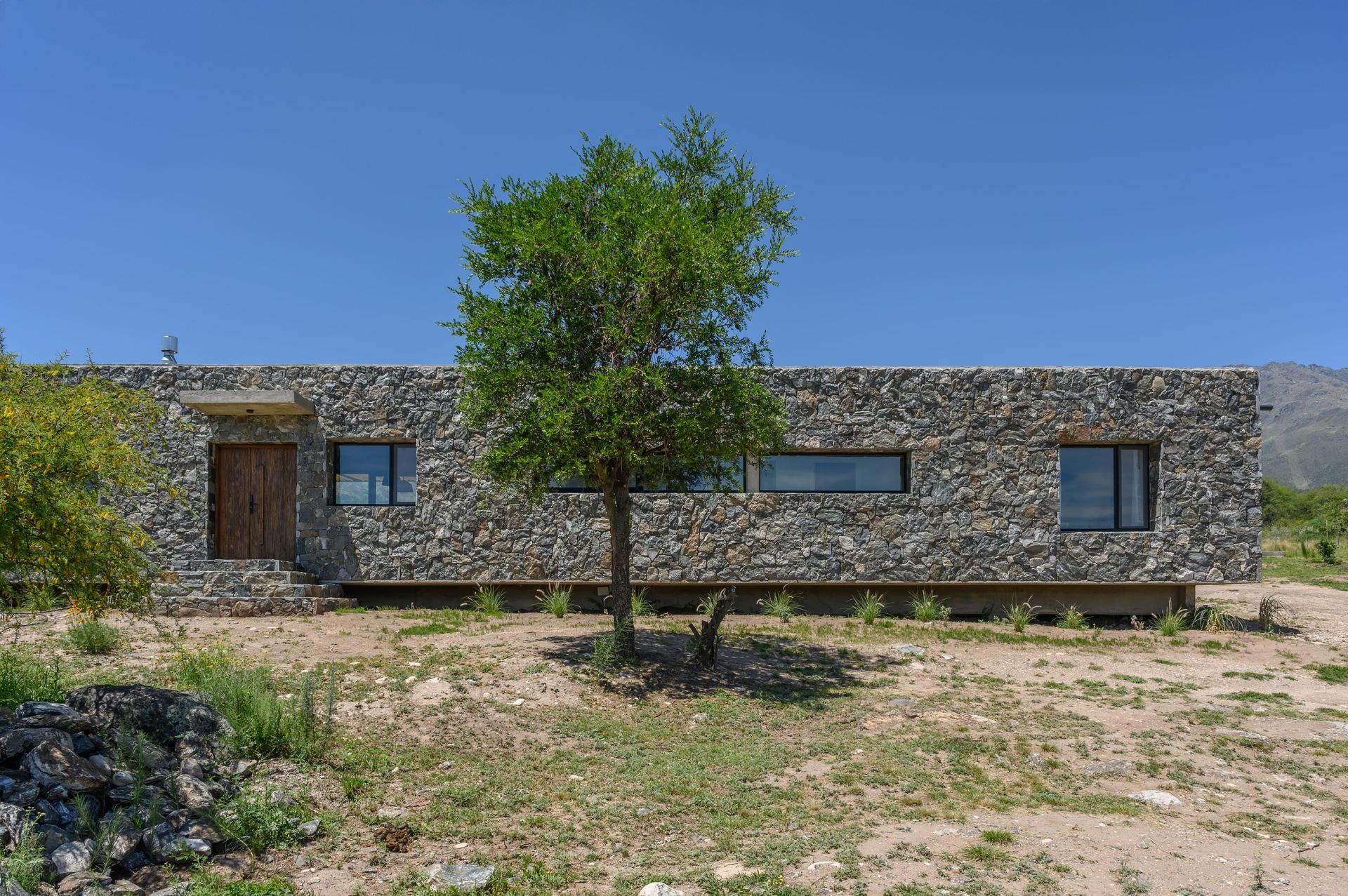 Casa rectangular revestida de piedra, con puerta y ventanas de madera, árbol al frente y contra un cielo azul.